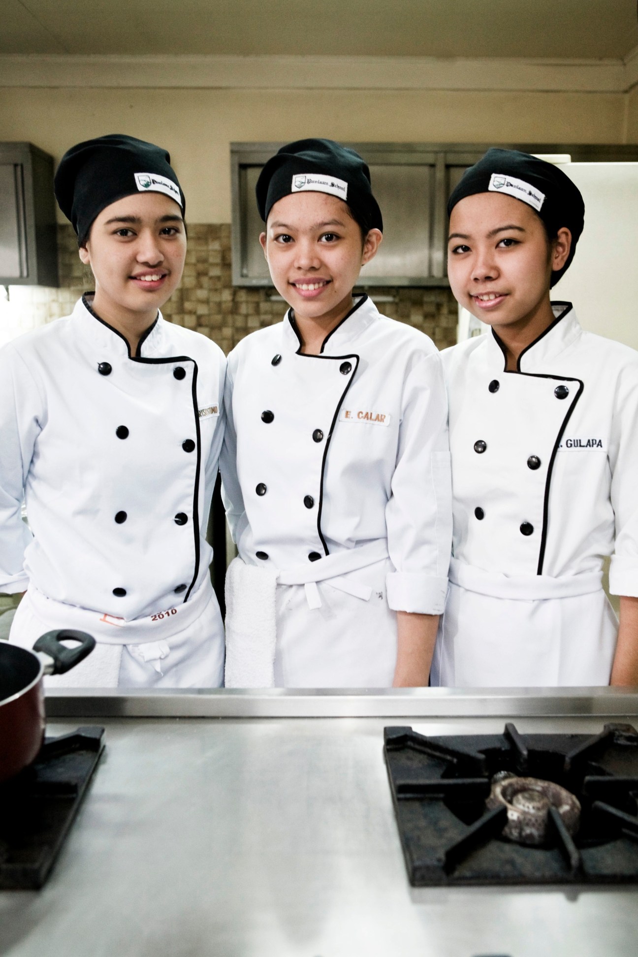 Three young female culinary students in white chef coats and headbands smile at Punlaan School kitchen.