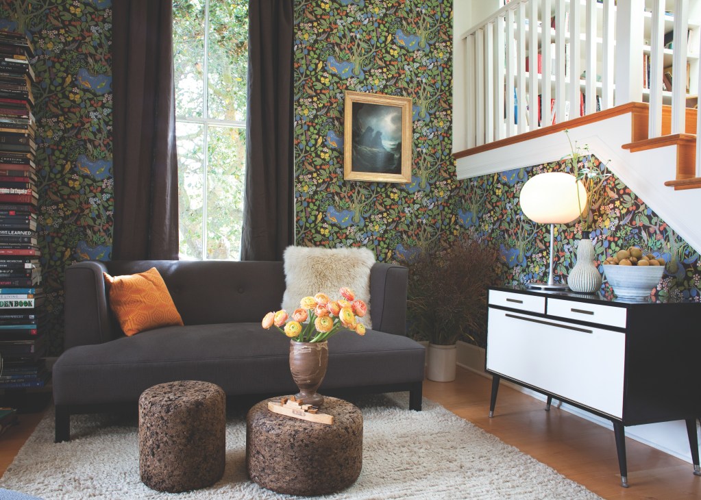 Living room with dark sofa, Josef Frank patterned wallpaper, white bookshelf, and orange flowers in ceramic vase.