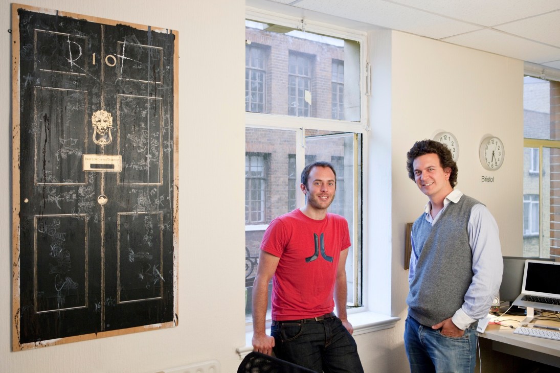 Two men stand in an office next to a large blackboard with the Riot logo and sketches on it.