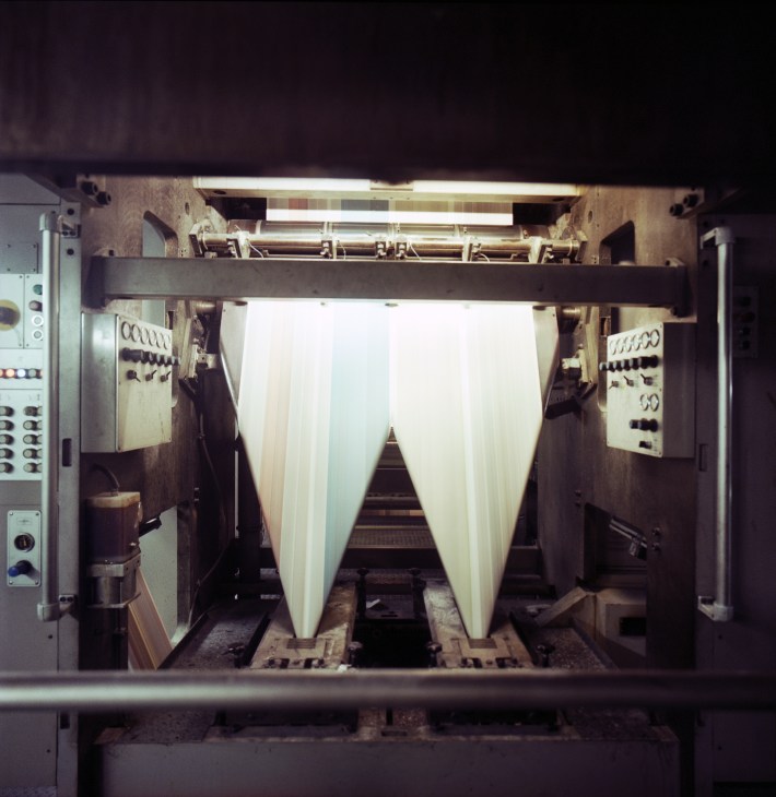 Industrial printing press machinery with large rolls of white paper feeding through the machine.
