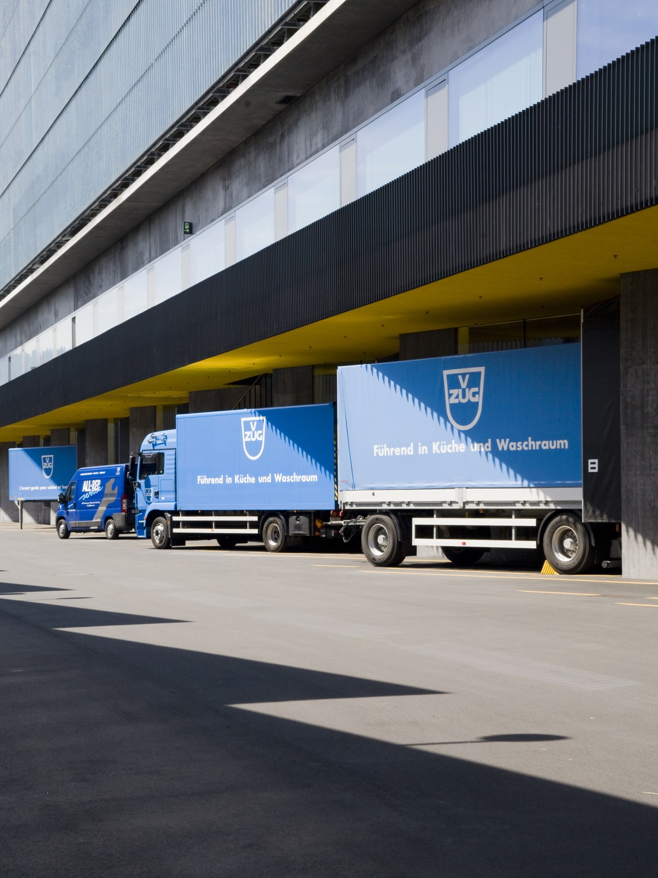 V-Zug branded blue delivery lorries parked outside an industrial logistics building with yellow and black striped canopy.