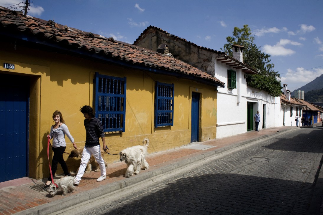People walk dogs on a cobblestone street lined with colonial buildings in bright yellow and white with blue doors and windows.