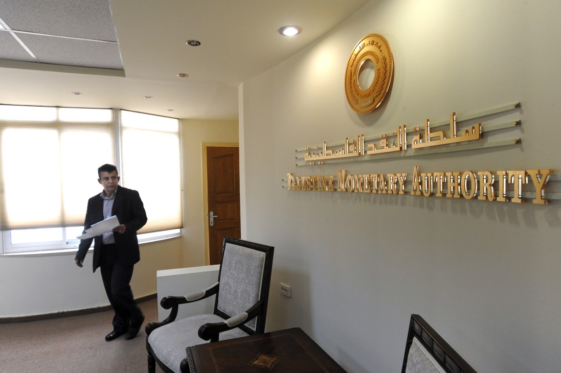 Man in dark suit holding papers in Palestine Monetary Authority office with gold signage on wall.