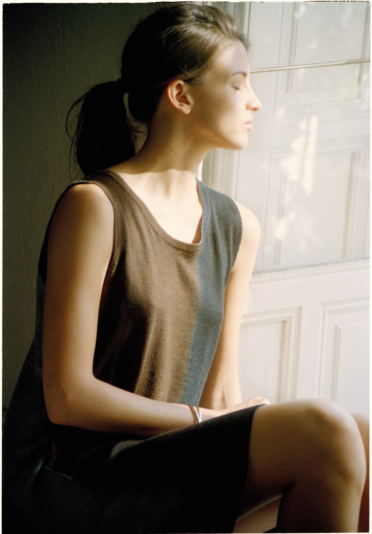 Model Carina Pichler wearing a grey sleeveless dress by C.P. Company and Cartier bracelet, seated by a window.