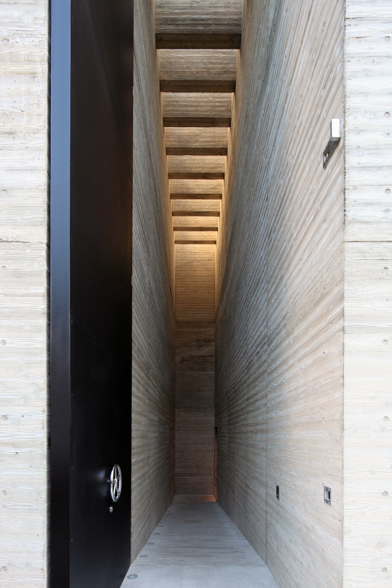 Narrow corridor with wooden ceiling slats and weathered wood walls flanking a dark door on the left.