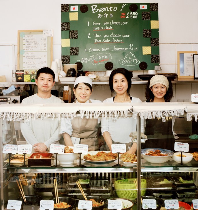 Four staff members stand behind a counter at Cha No Ma, a Japanese café in London, with a handwritten bento menu board displayed above.