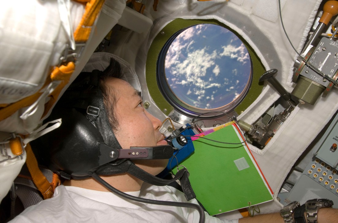 Astronaut Koichi Wakata looks through a window on the Soyuz TMA-14 spacecraft, with Earth visible beyond the porthole.