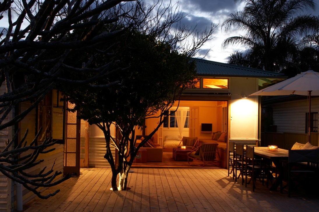 Evening view of a modern beach house with illuminated interior, wooden deck, and outdoor dining area surrounded by trees.