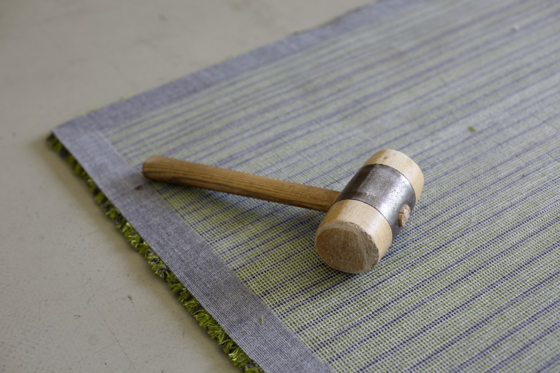 A wooden hammer with metal band rests on a blue and green striped carpet sample.
