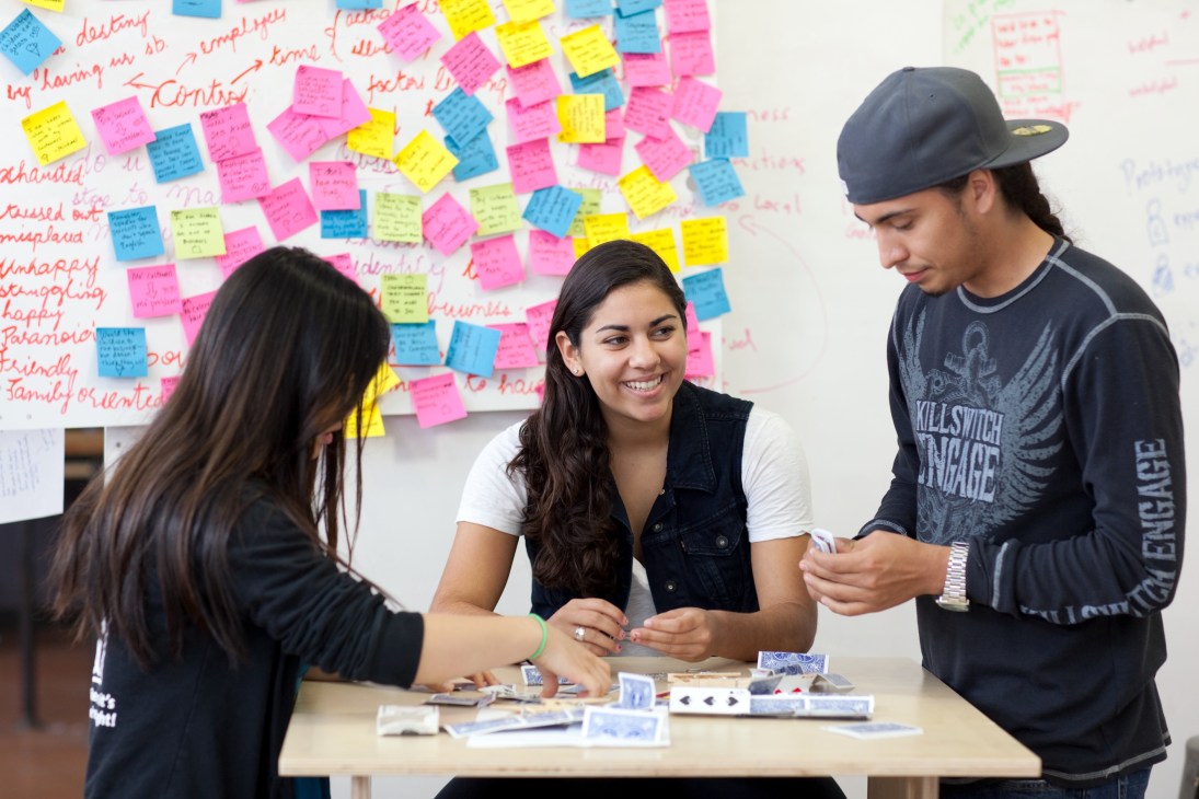 Three d.school students collaborating at a table with design materials, with colorful sticky notes on a whiteboard behind them.