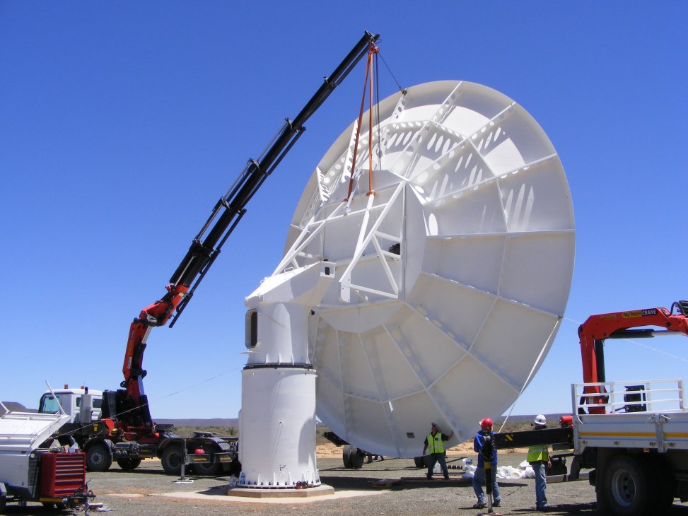 Construction workers install a large white satellite dish antenna using a red crane in a desert landscape under clear blue sky.