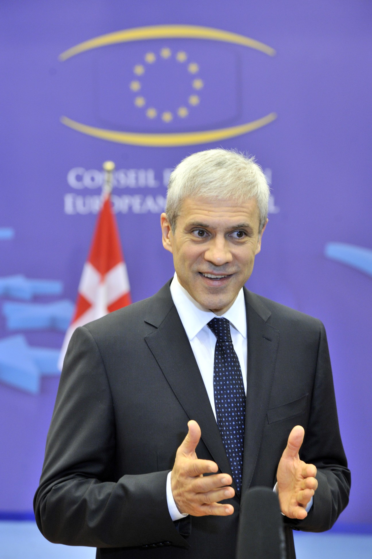 Man in dark suit and blue tie speaking at podium with EU flag and Council of Europe logo in background.