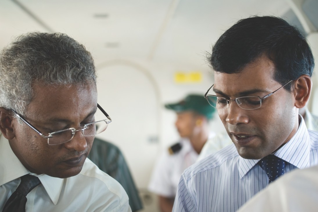 Two men in business attire review documents together indoors, with other people visible in the background.