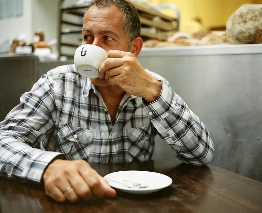 Nadav Kander drinks coffee from a white cup in a kitchen setting with bread visible in the background.