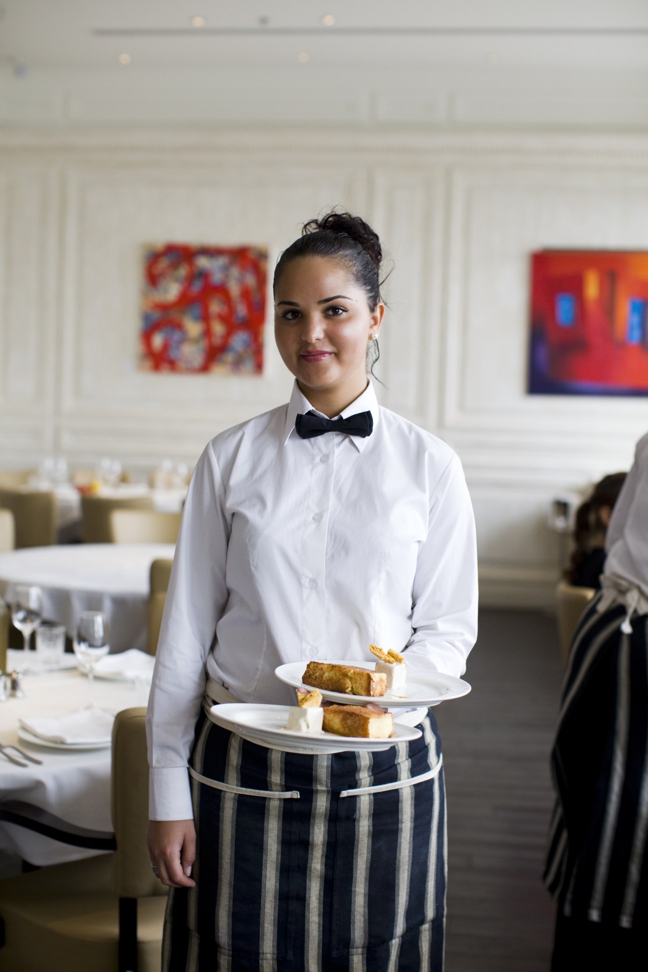 Waiter in white shirt and bow tie holding plates of food in an upscale restaurant dining room.