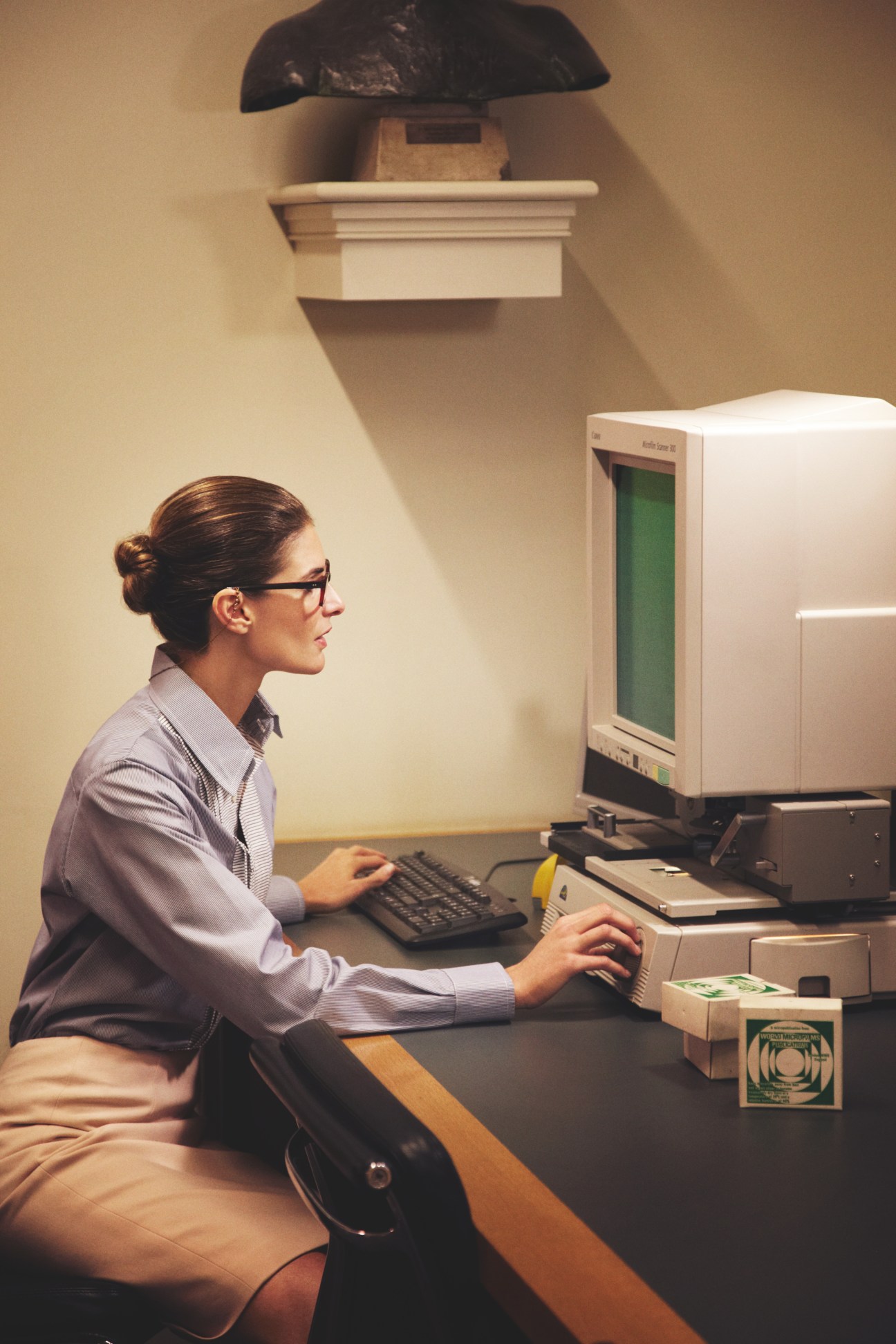 Woman wearing glasses and business attire at a retro computer terminal in a library setting.