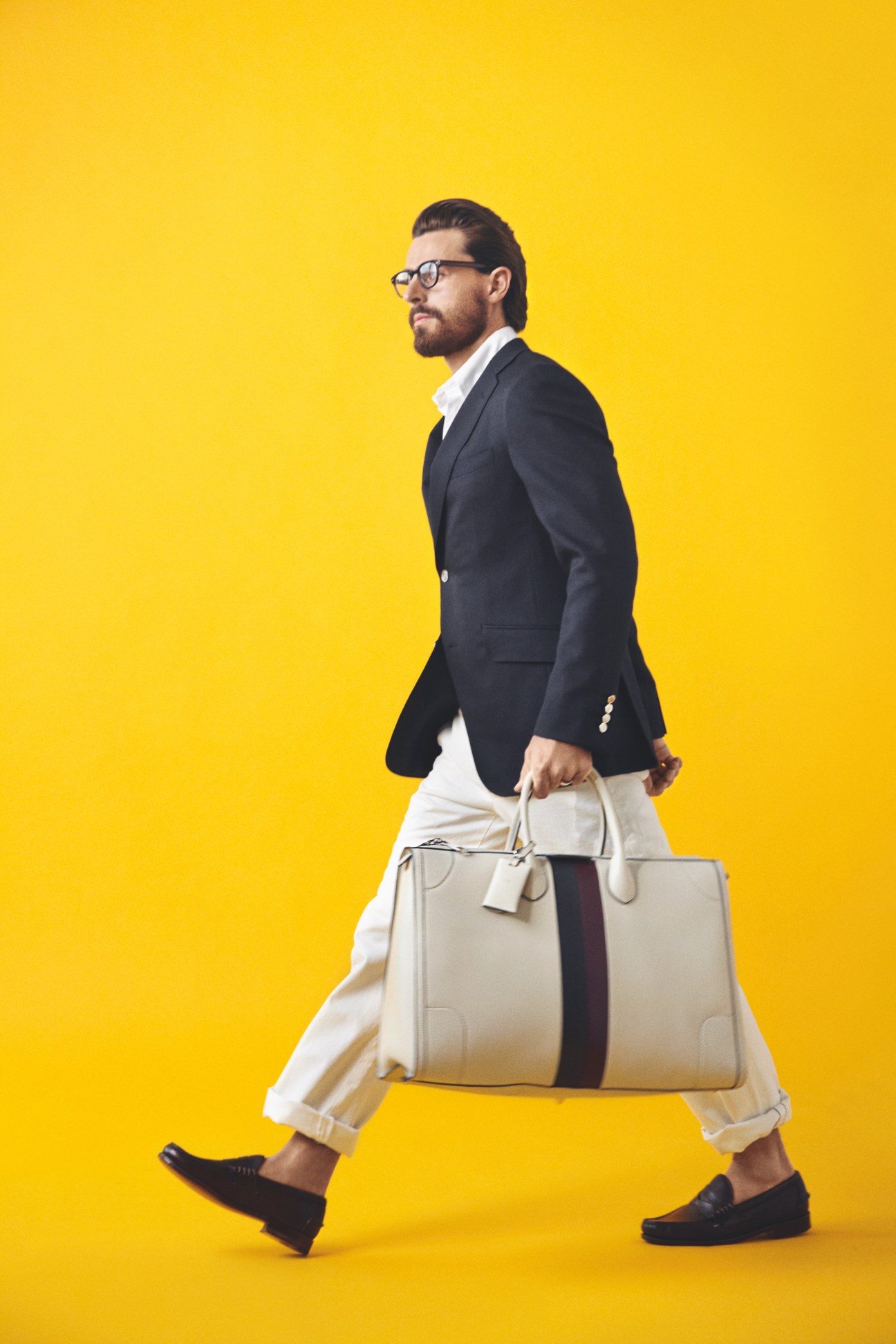 Man in black Dunhill jacket and white Todd Snyder trousers carrying a Gucci bag against yellow background.