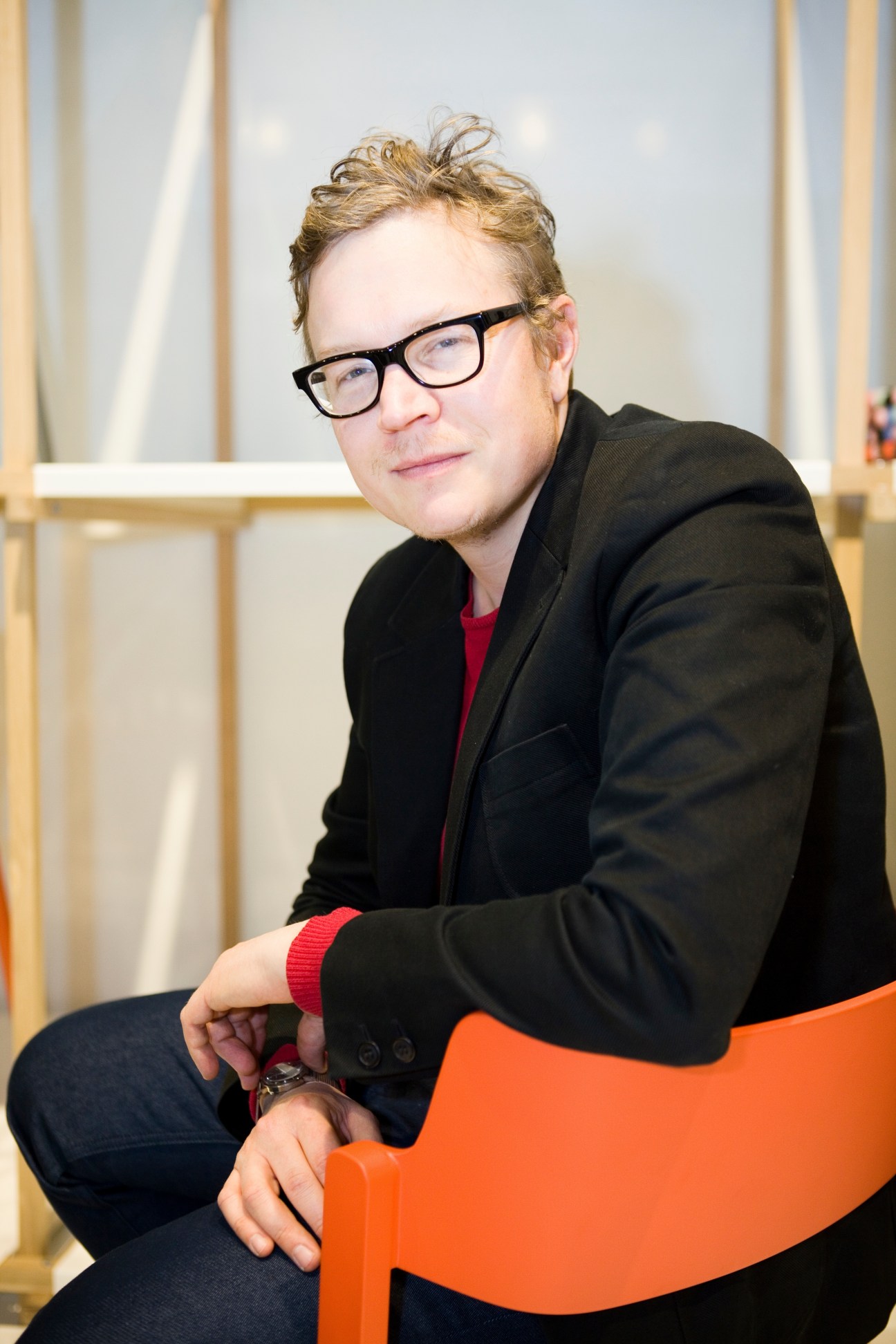 Designer Shane Schneck sits on an orange chair wearing black-framed glasses and a black blazer.