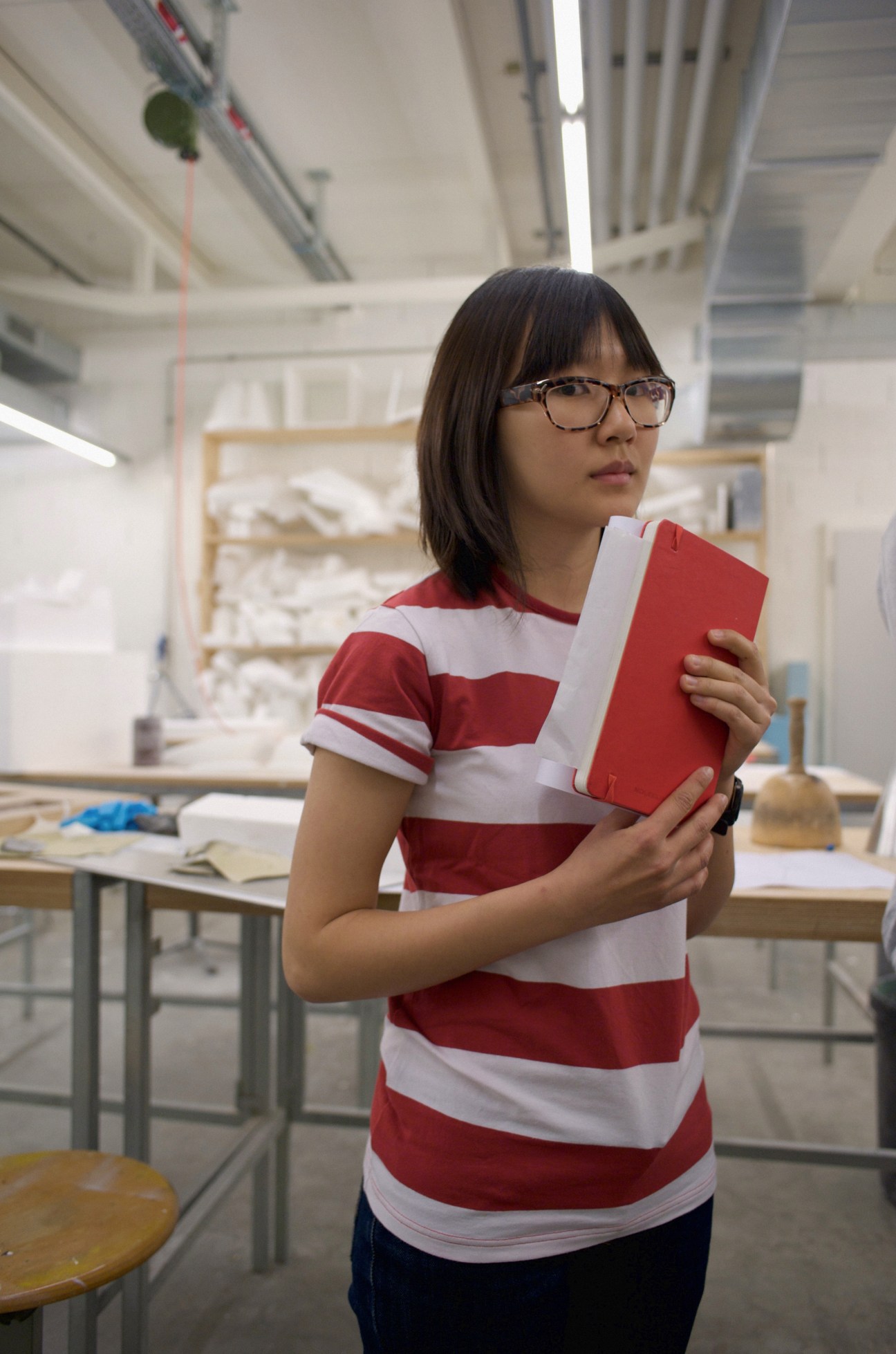 A student wearing glasses and a red-and-white striped shirt holds a red notebook in an ECAL workshop.