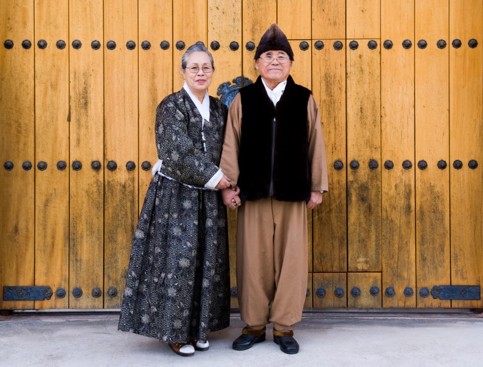 Lee Bong-ju and Sa Jun-ja stand hand-in-hand wearing traditional Korean formal dress against a wooden door with metal studs.