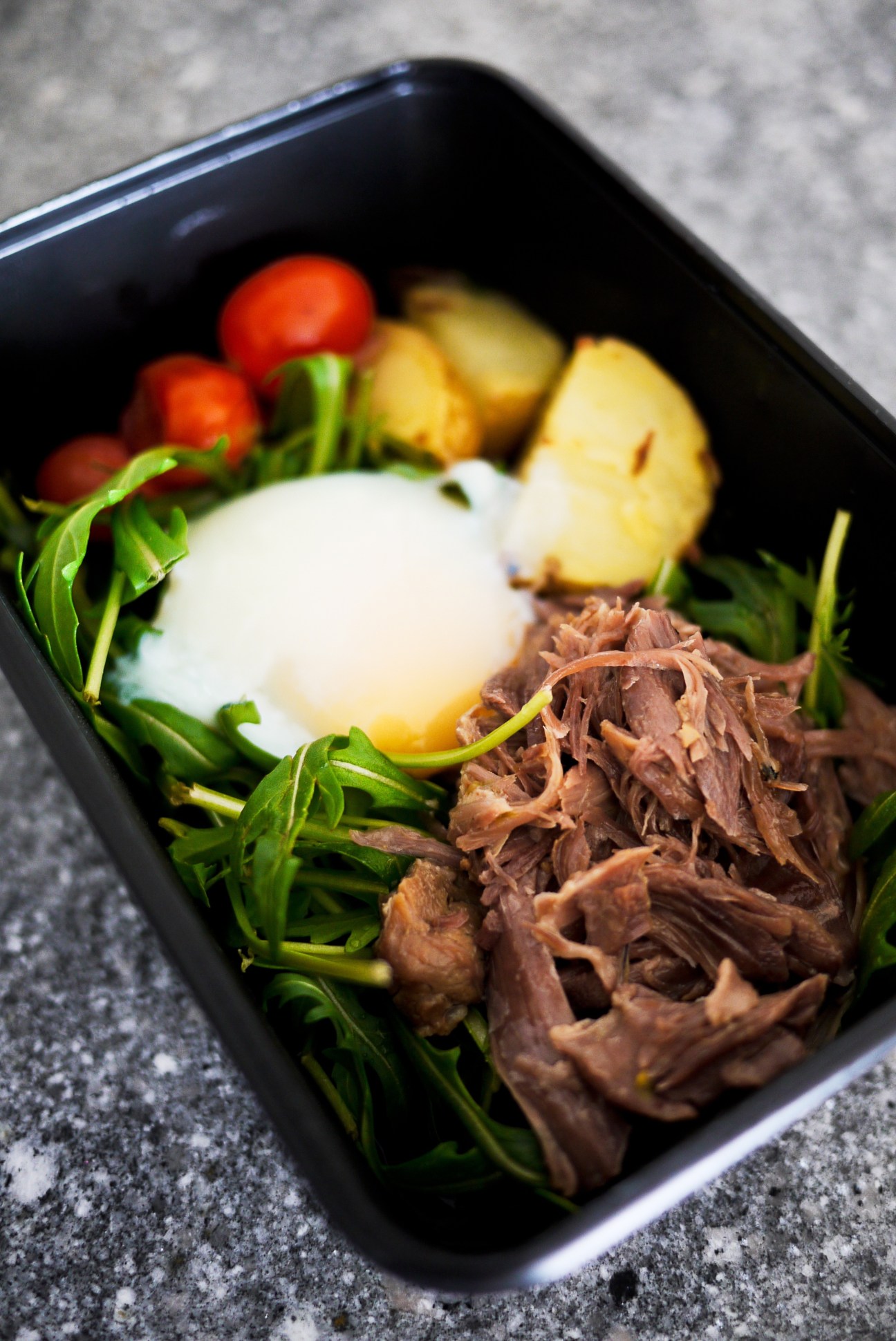A black container filled with shredded meat, green vegetables, potatoes, tomatoes and a white sauce dollop.