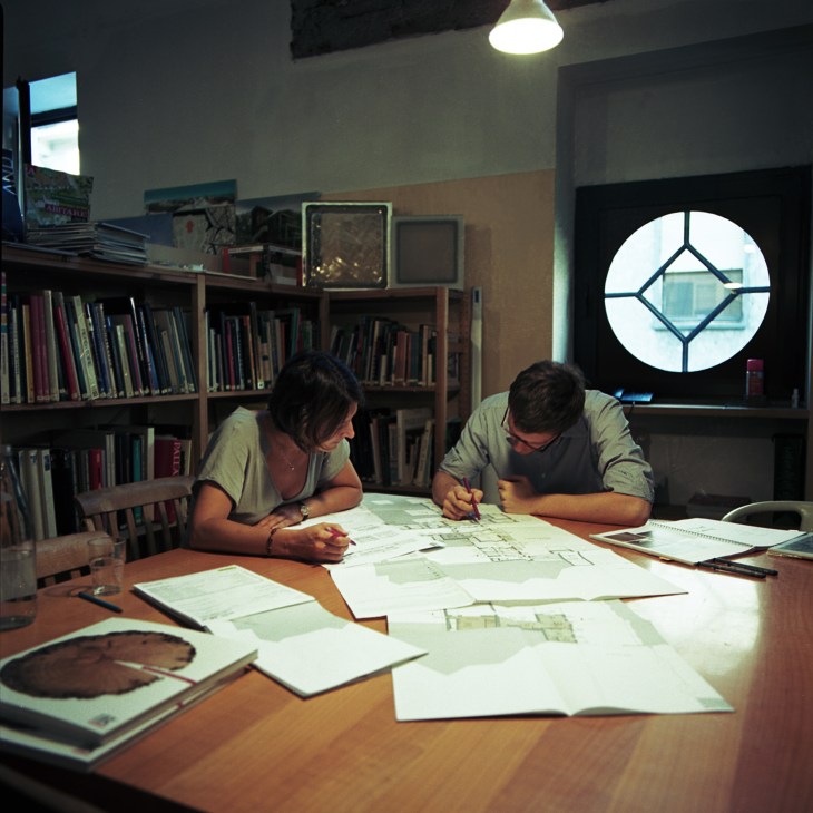 Two staff members examine architectural blueprints and drawings at a wooden table in a library-filled office space.