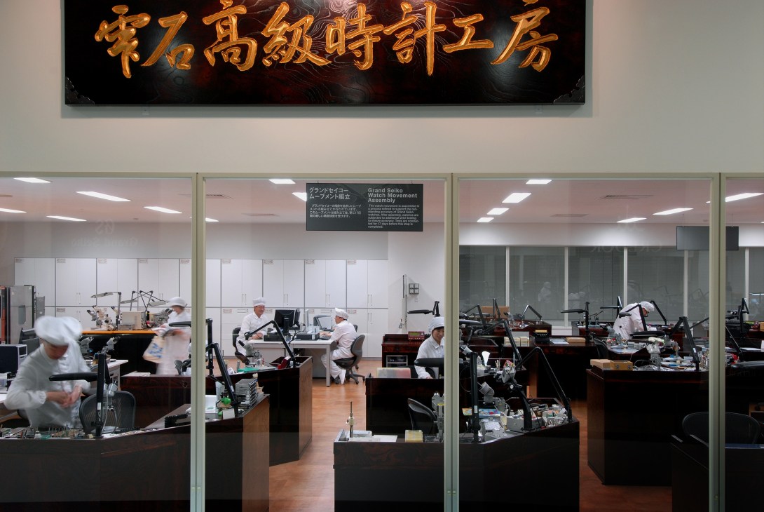 Watch factory workers in white uniforms assemble timepieces at workstations behind glass walls in the Jura Mountains.