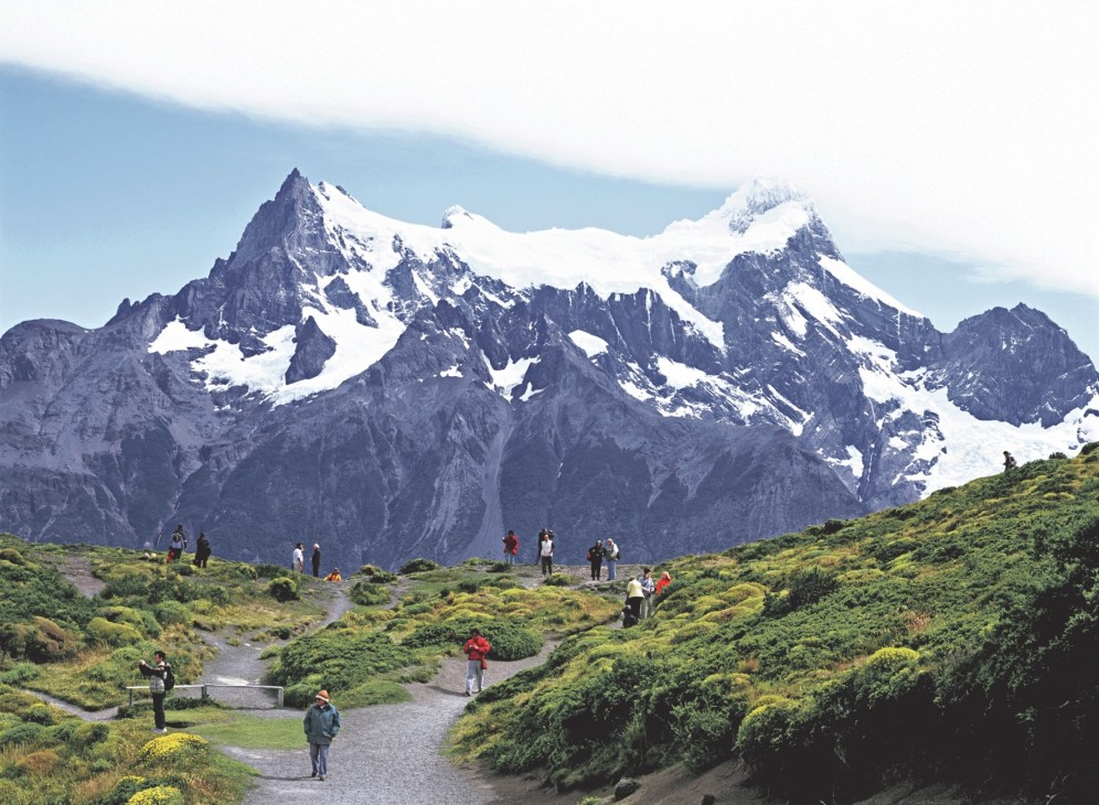 Snow-capped mountain peaks of Torres del Paine with hikers on green valley paths below.