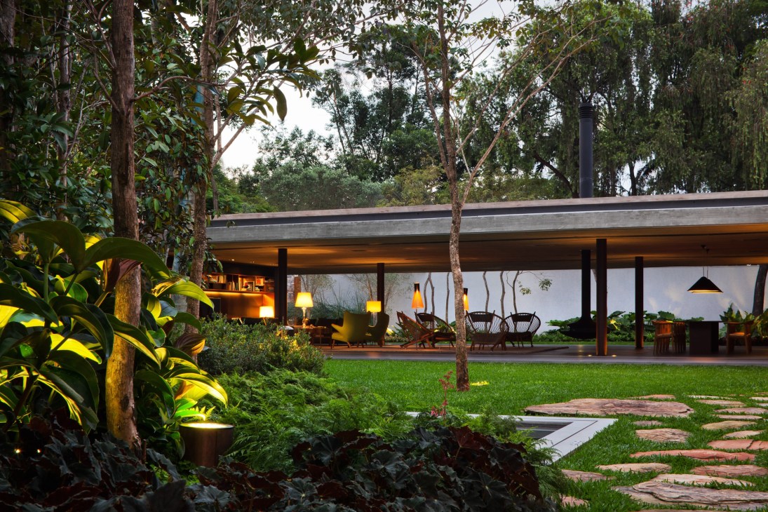 Evening view of V4 residence's glass-walled pavilion lit from within, surrounded by tall trees and manicured garden in São Paulo.