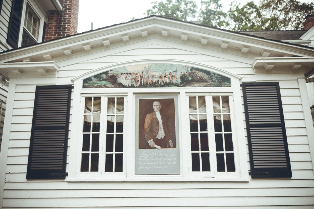 Roger Sherman Inn's white clapboard exterior facade featuring a portrait and memorial of Roger Sherman between windows with black shutters.