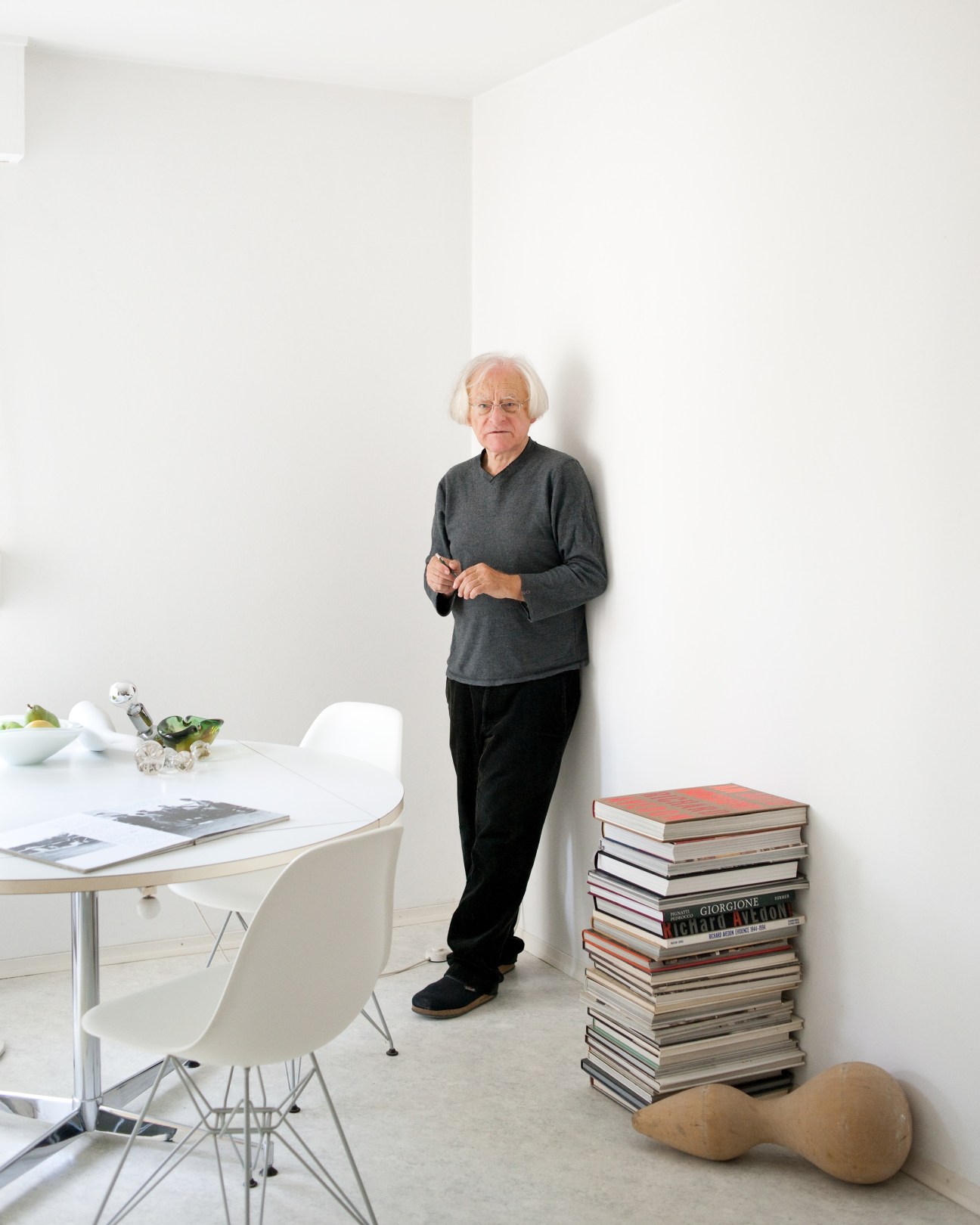 Jürgen Thiele, an elderly man with white hair wearing a dark sweater, stands in his minimalist apartment next to stacked books and a round table.