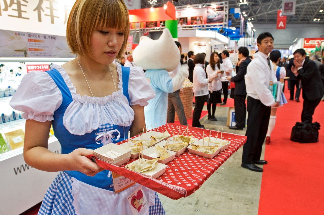 A woman in a blue and white costume dress holds a tray of food samples at a trade show exhibition hall.