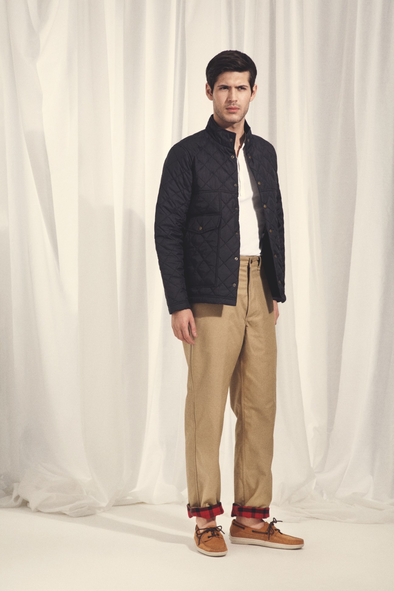 Man wearing black quilted jacket, white shirt, khaki trousers with plaid cuff, and brown shoes against white curtain backdrop.