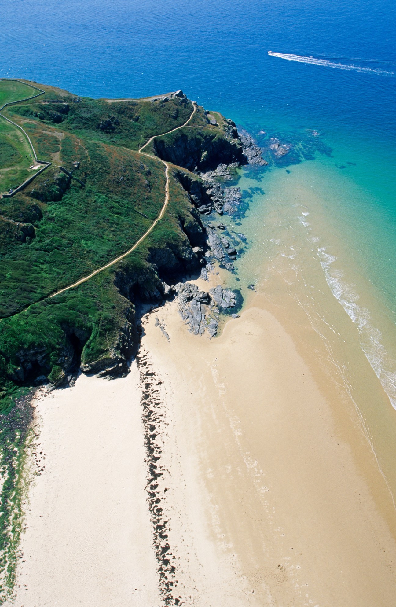 Aerial view of a green coastal headland with rocky outcrops meeting a sandy beach and turquoise ocean.