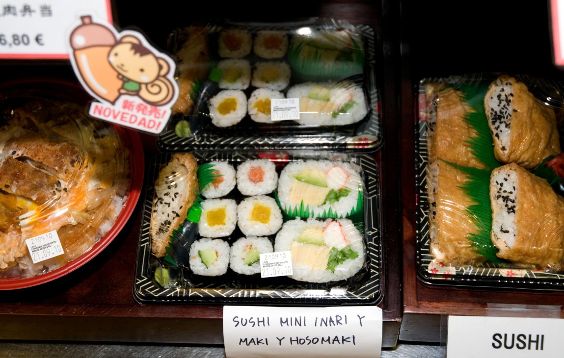 Sushi and Japanese prepared foods displayed in plastic containers in a shop window with price labels and a cartoon character sign.