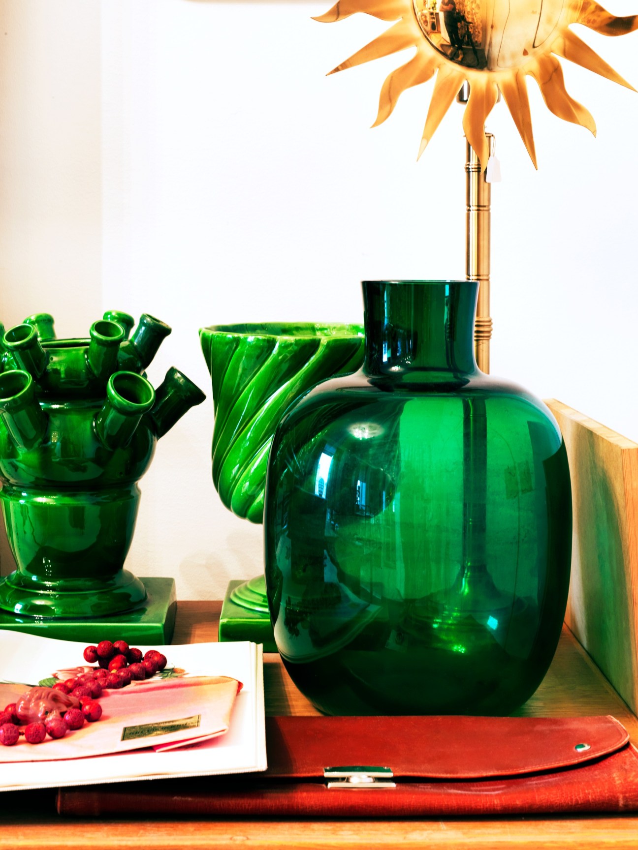 Green glass vessels and a gold sunburst sculpture displayed with red raspberries and accessories on a wooden desk.
