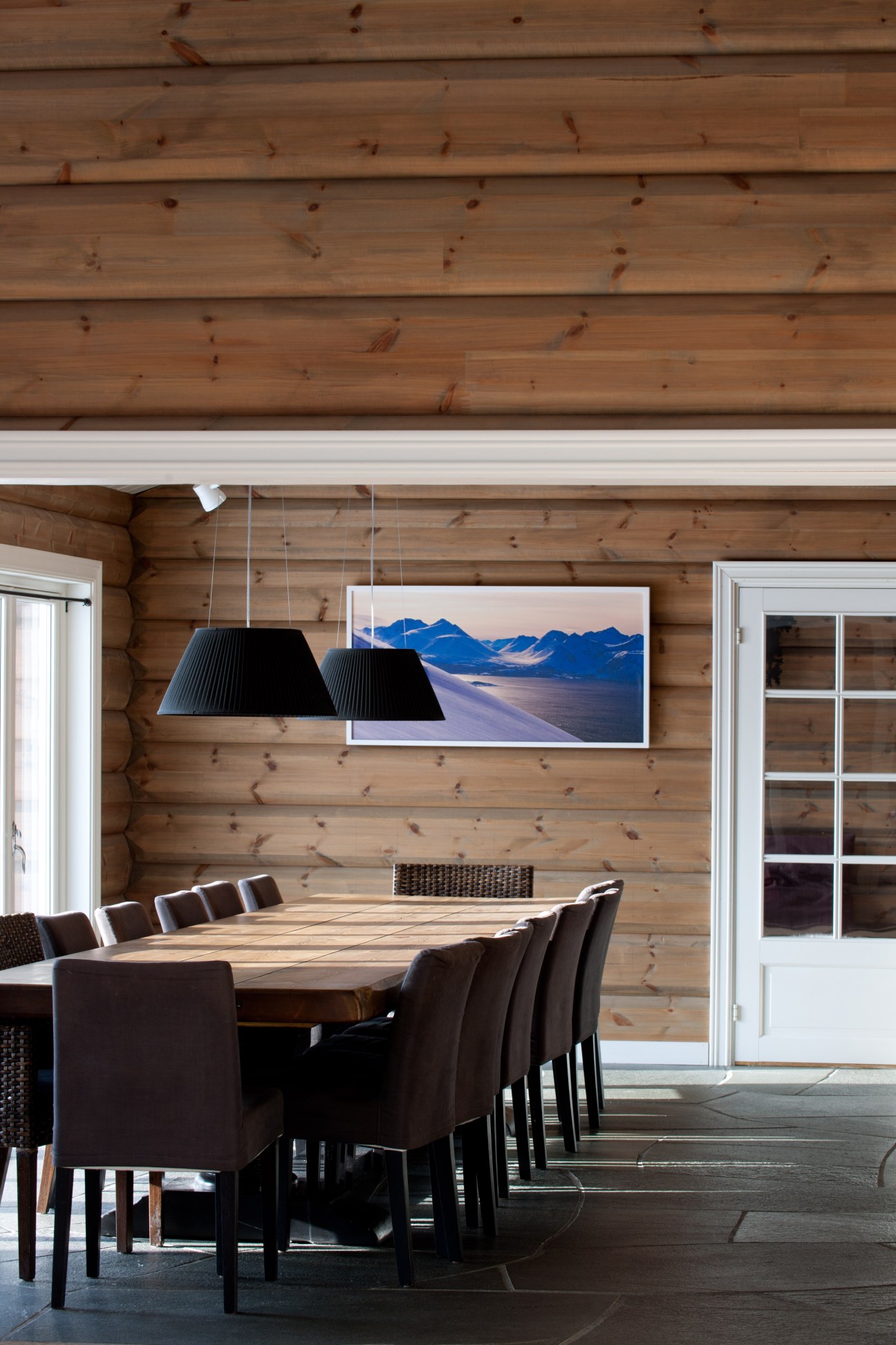 Modern dining room with log cabin walls, long wooden table, brown chairs, black pendant lights, and mountain landscape artwork.