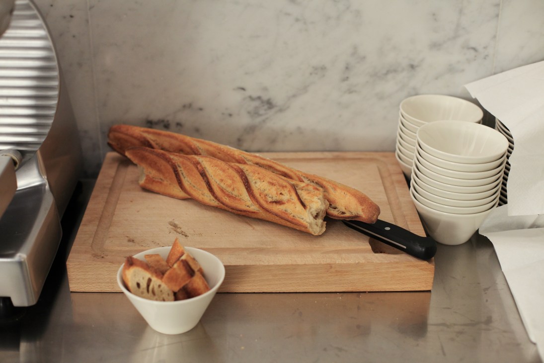 A sliced baguette on a wooden cutting board with white bowls and a marble countertop at Le Dauphin restaurant.