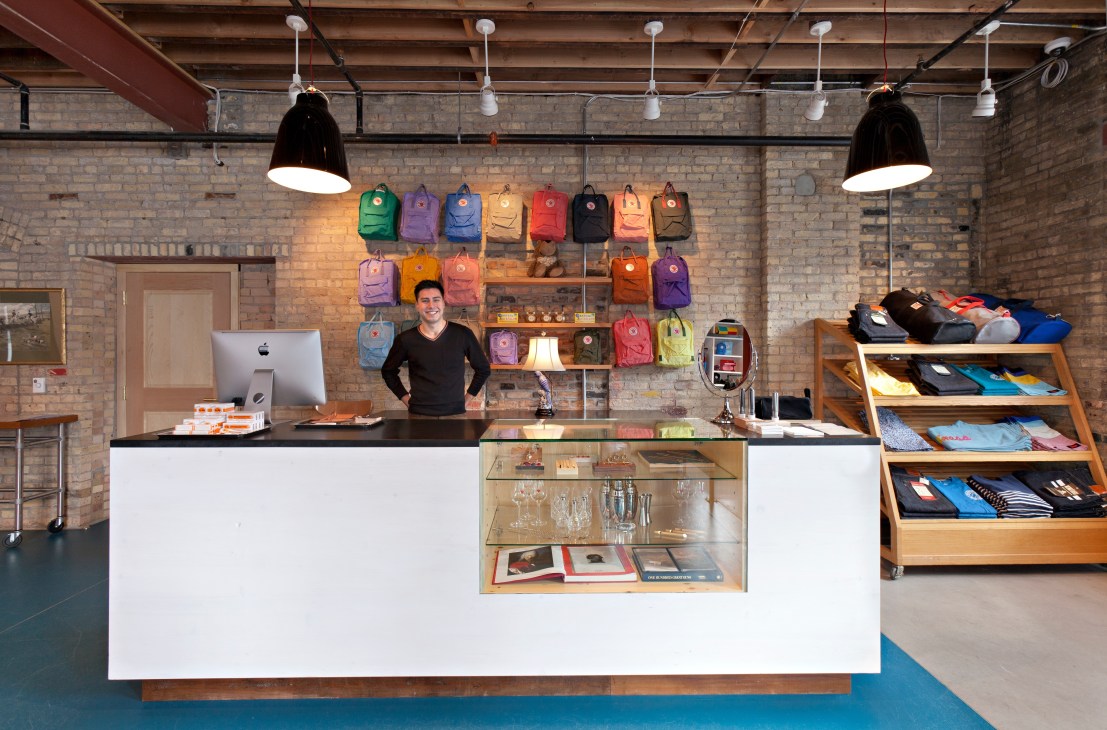 Interior of Askov Finlayson retail store with colorful backpacks displayed on brick wall and staff member at counter.