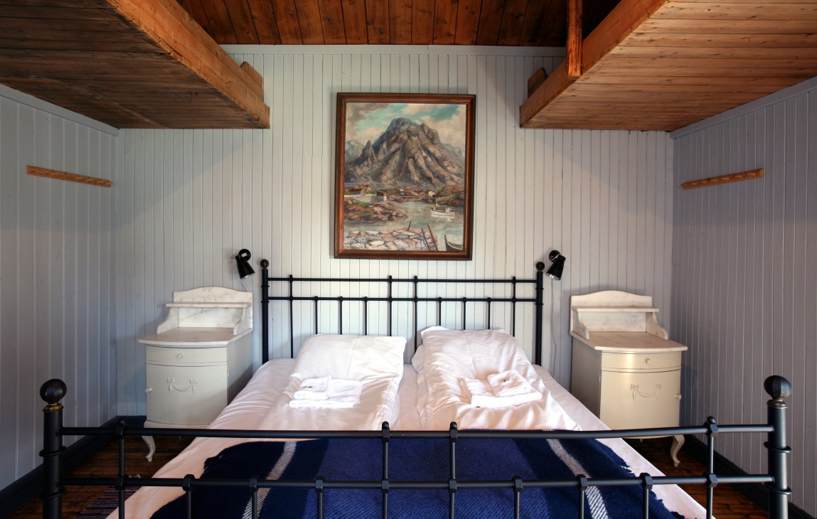 Interior of a Reine Rorbuer cabin bedroom with black metal bed frame, white bedding, and wooden ceiling beams.