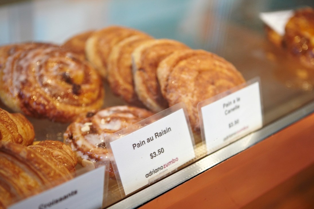 Pastries displayed in a bakery case with price labels, including a Pain au Raisin labeled $3.50 at Adriano Zumbo.