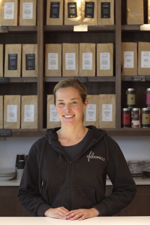 Woman in Fibonacci bakery hoodie standing at counter with shelves of packaged coffee beans behind her.