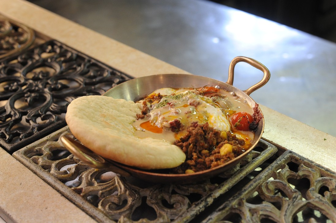 A brass bowl with handles containing a flatbread and meat dish with vegetables on an ornate metal trivet.