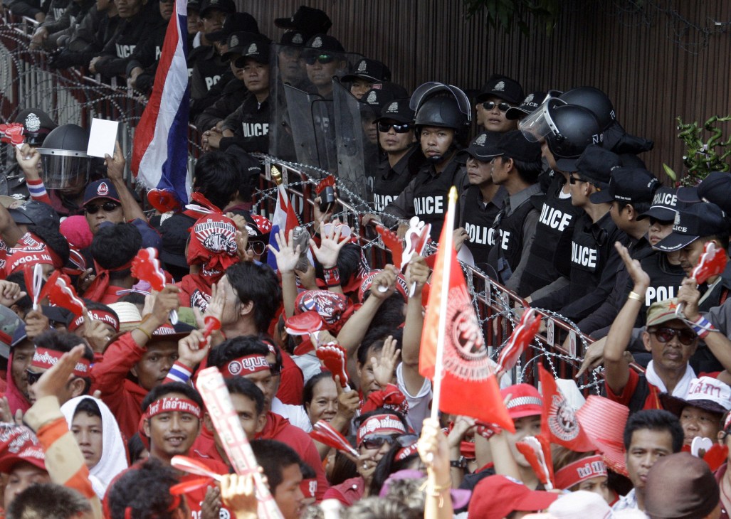 Thai police in riot gear stand behind barbed wire facing Red Shirt protesters holding Thai flags.