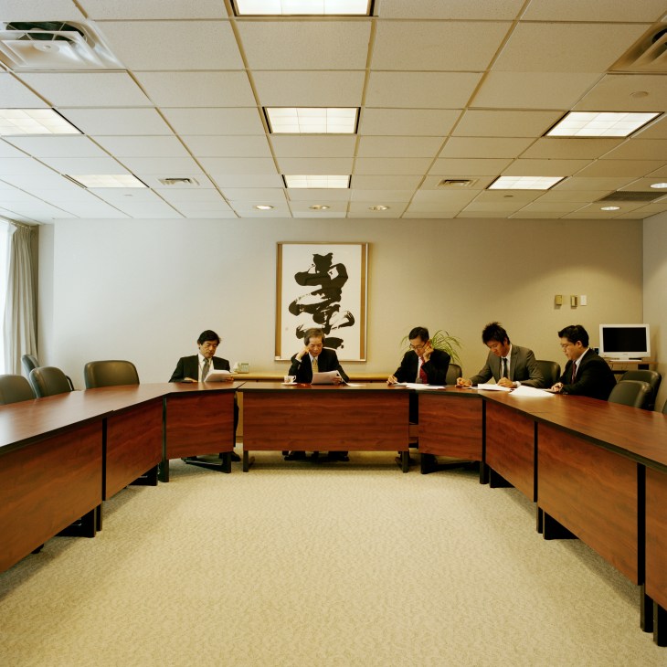 Japanese diplomats seated at a U-shaped wooden table during a staff meeting in a modern conference room.