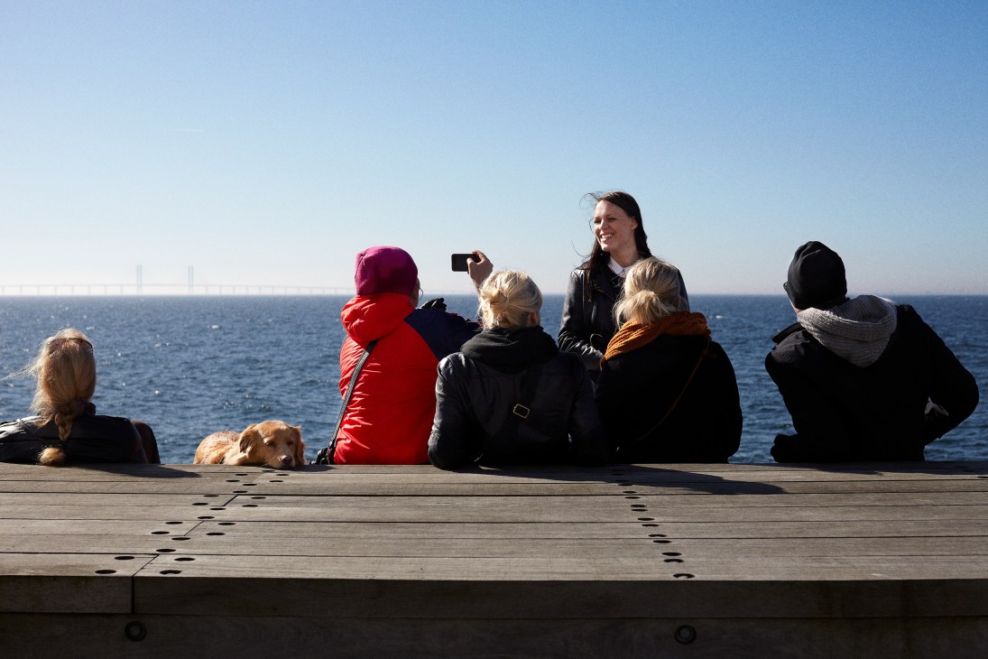 People and a dog sitting on a wooden promenade facing the sea at Sundspromenaden, Västra Hamnen in Malmö.