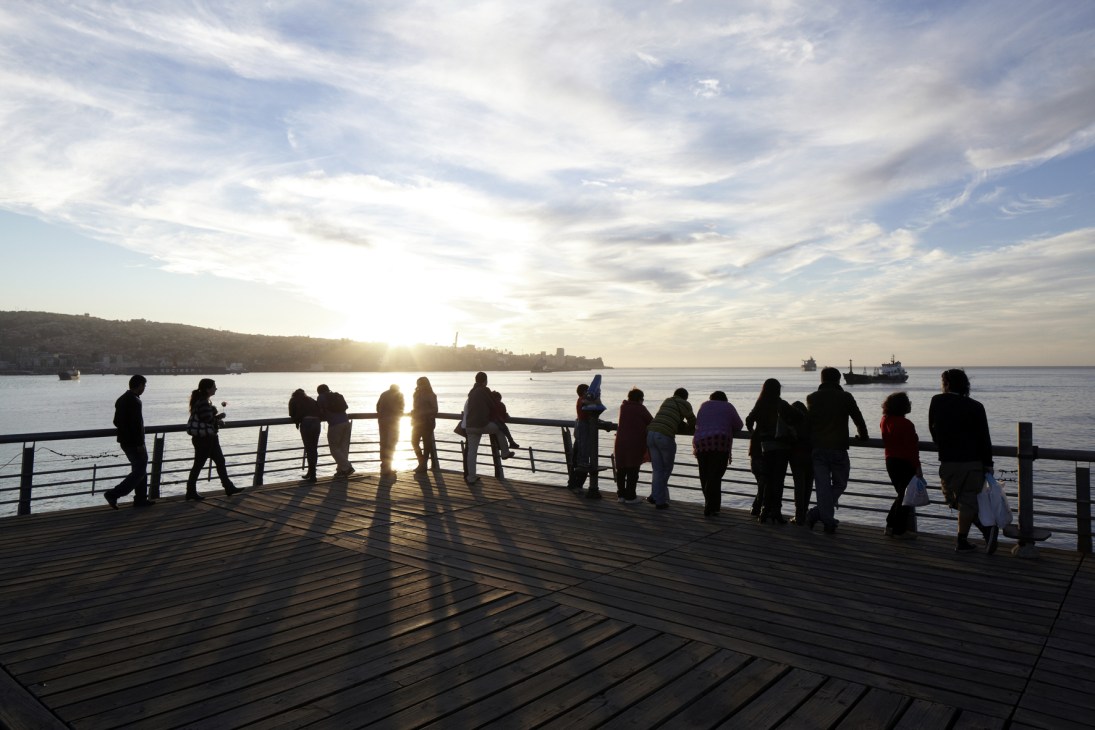 Silhouetted visitors stand along a wooden pier railing at sunset overlooking Valparaíso's Pacific harbour.