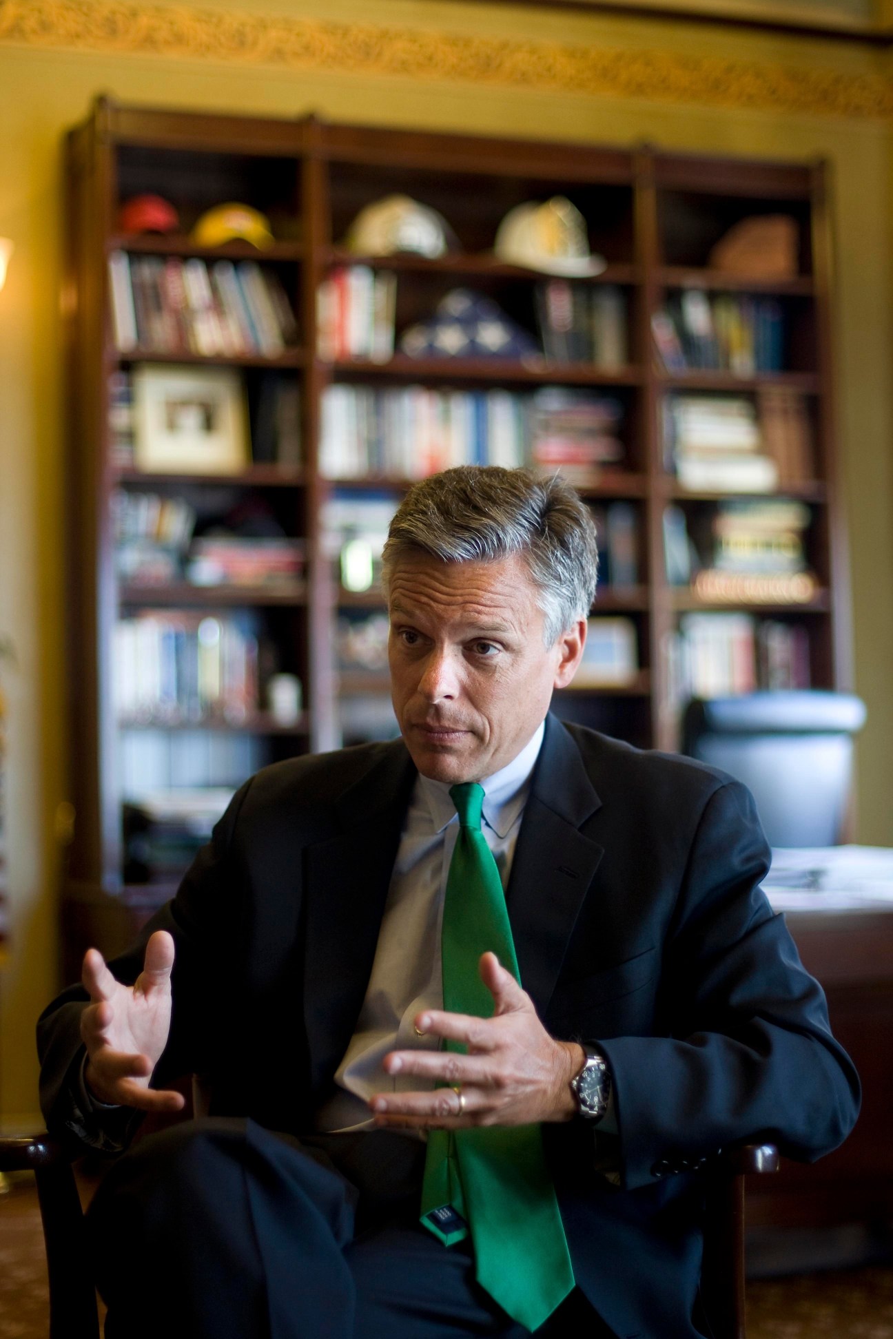 Man in business suit with green tie gesturing while speaking in front of a bookshelf.