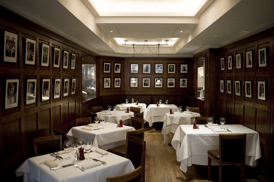 Classic wood-panelled dining room at J Sheekey restaurant with white-clothed tables and framed photographs on walls.