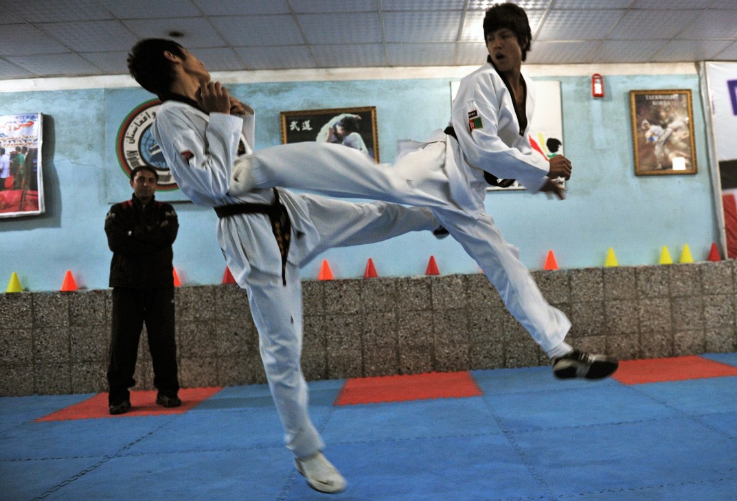 A taekwondo athlete in white uniform performs a high kick during sparring practice while coach watches from the sideline.
